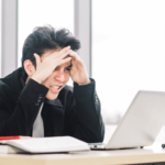 A stressed businessman in a black suit sitting at a desk, holding his head in his hands with a laptop and papers including a QA campaign report in front of him, displaying frustration or headache in a bright