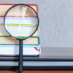 A magnifying glass in front of a stack of books and a closed laptop on a wooden desk, primed for search results analysis.